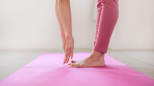 Person in rosa Leggings steht auf einer pinken Yogamatte und beugt sich mit ausgestreckter Hand nach unten.