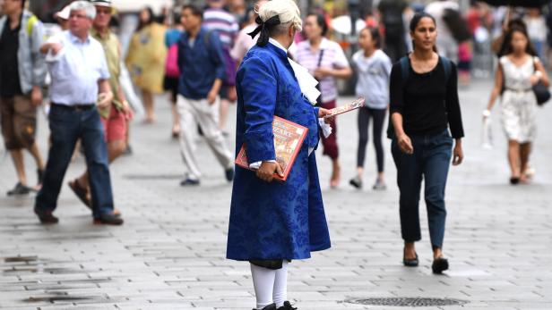 Ein Mann im blauen Barockkostüm verteilt Flugblätter auf einer belebten nassen Straße.