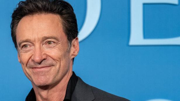 Hugh Jackman lächelt vor einem blauen Hintergrund in einem grauen Sakko und einem schwarzen Hemd.