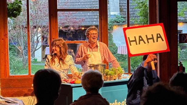 Zwei Menschen kochen vor Publikum, während eine Person ein „HAHA“-Schild hochhält.