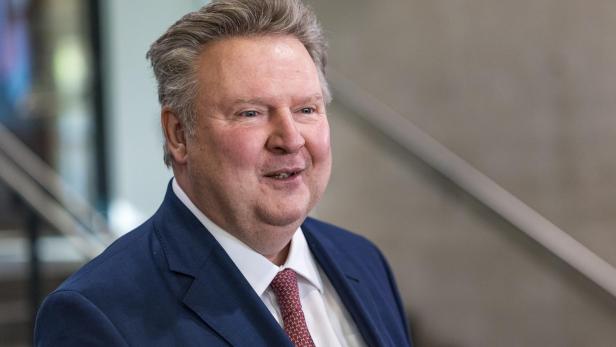 Ein lächelnder Mann im blauen Anzug mit roter Krawatte vor neutralem Innenraumhintergrund.