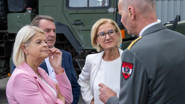 Vier Personen sprechen vor einem Militärfahrzeug, darunter eine Frau in Pink und ein Offizier in Uniform.