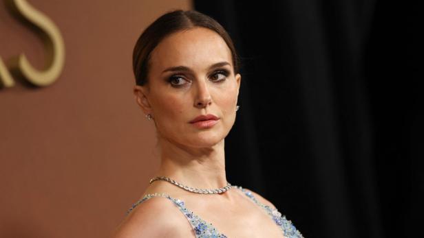 Natalie Portman trägt ein mit kleinen blauen Blumen verziertes Kleid und eine dezente Halskette vor einem dunklen Hintergrund.