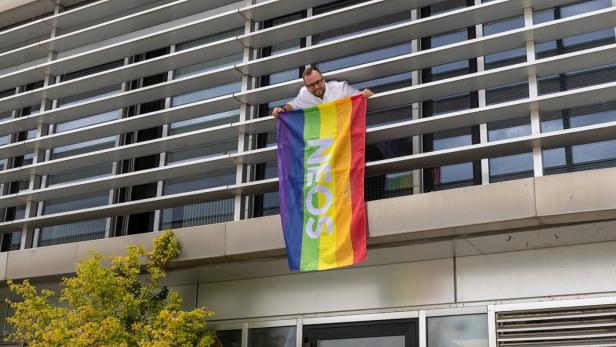 Eine Person hängt auf einem Balkon eine Regenbogenflagge mit „NEIN“ an ein modernes Gebäude.