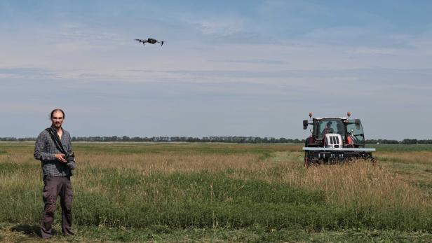 Mann steuert eine Drohne über einem Feld, neben einem Traktor.