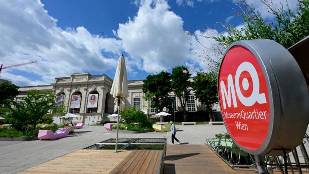 MuseumsQuartier Wien mit großem rotem MQ-Schild, Platz, Bäumen, Wolkenhimmel und historischer Fassade.