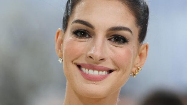 Anne Hathaway mit dunklem Haar, großem goldenen Collier und trägerlosem schwarz-weißem Kleid vor unscharfem Hintergrund.
