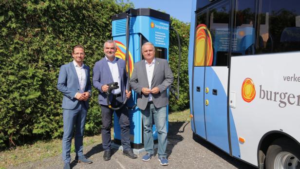 Drei Männer stehen neben einem Linienbus und einer blauen Ladesäule für Elektrofahrzeuge.