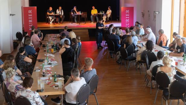 Publikum sitzt bei einer Podiumsdiskussion in einem Saal mit Bühne und roten KURIER-Bannern.