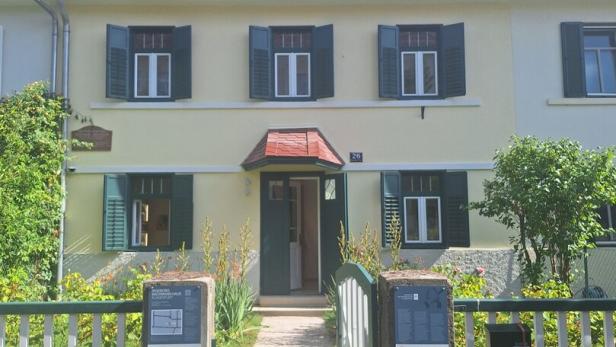 Das Ingeborg-Bachmann-Haus in Klagenfurt ist ein hellgelbes Gebäude mit rotem Ziegeldach, grünen Fensterläden und einem kleinen Vorgarten.