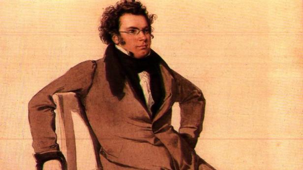Ein Mann mit Locken, Brille und braunem Mantel sitzt lässig auf einem Stuhl vor hellem Hintergrund.