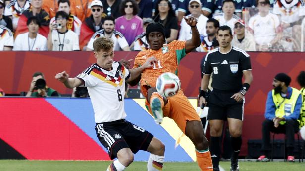Ein deutscher Spieler im weißen Trikot und ein ivorischer Spieler in Orange kämpfen auf dem Rasen um den Ball, beobachtet von einem Schiedsrichter.