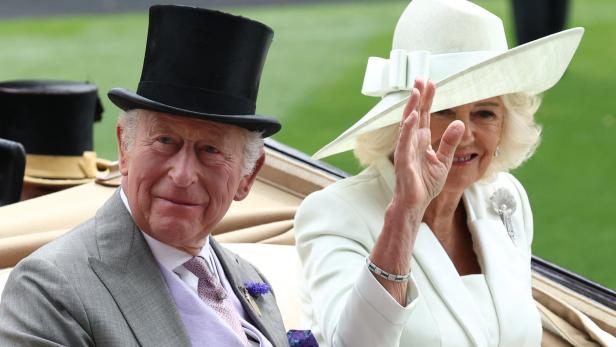 König Charles III. und Königin Camilla winken in festlicher Kleidung aus einer Kutsche.