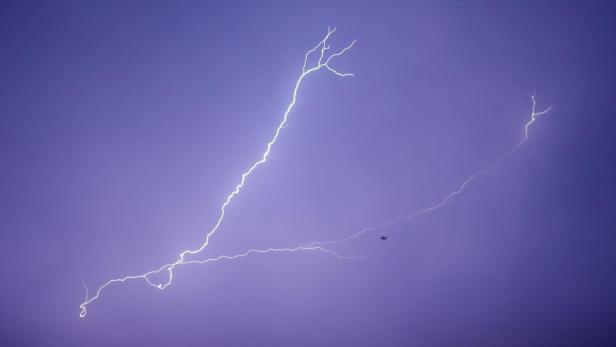 Gewitterhimmel mit mehreren hellen Blitzen und einem kleinen Flugzeug in der Ferne.