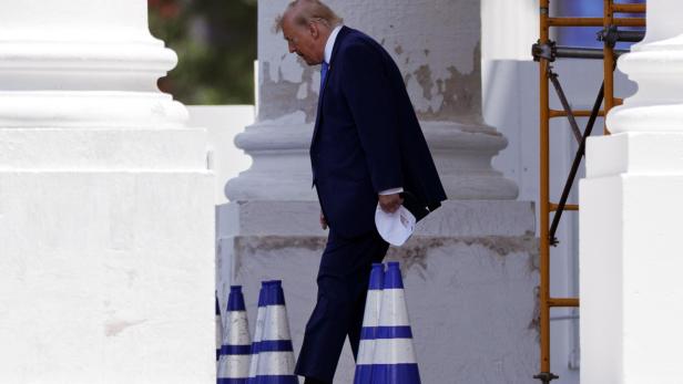Donald Trump geht mit gesenktem Kopf an weißen Säulen und blau-weißen Pylonen vorbei.