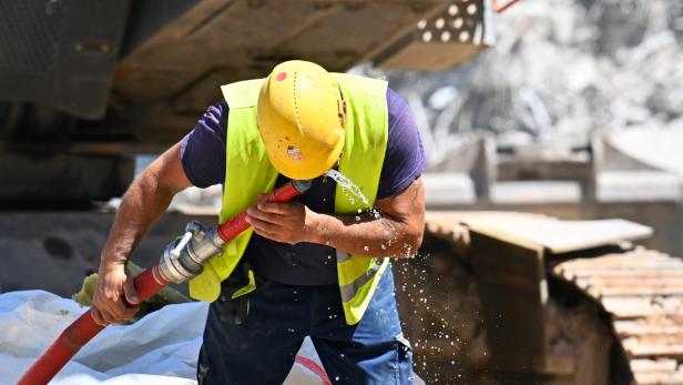 Ein Bauarbeiter mit gelbem Helm und Warnweste trinkt Wasser aus einem roten Schlauch auf einer Baustelle.