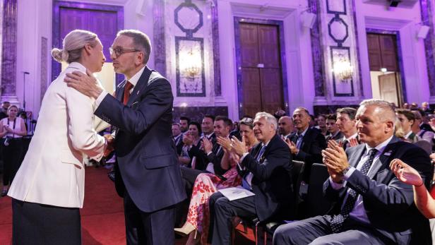 Alice Weidel und ein Mann in Anzug begrüßen sich herzlich vor einem applaudierenden Publikum in einem festlichen Saal.