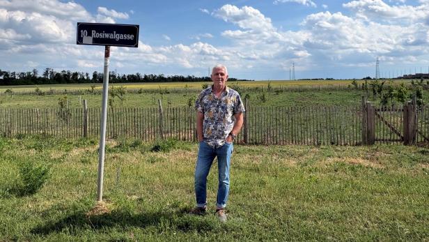 Mann steht neben dem Straßenschild „10. Rosiwalgasse“ auf einer Wiese vor Feldern unter wolkigem Himmel.