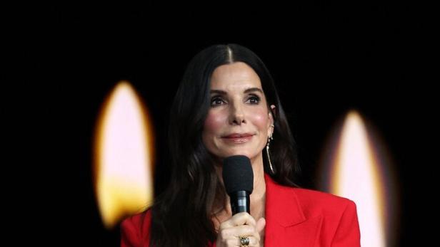 Sandra Bullock spricht in einem roten Hosenanzug vor einem schwarzen Hintergrund mit unscharfen Kerzenflammen in ein Mikrofon.