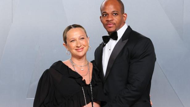Jennifer Meyer in einem schwarzen Oberteil und Geoff Ogunlesi im Smoking stehen eng beieinander vor einem hellgrauen Hintergrund.