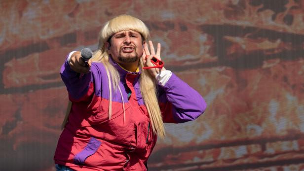 Oliver Tree mit blonder Vokuhila-Perücke und bunter Windjacke hält ein Mikrofon in die Menge und hält sich die Hand ans Ohr.