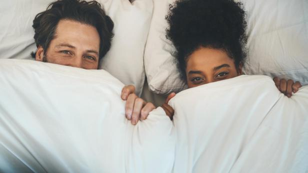 Ein lächelndes Paar liegt nebeneinander im Bett und schaut aus weißen Decken hervor.