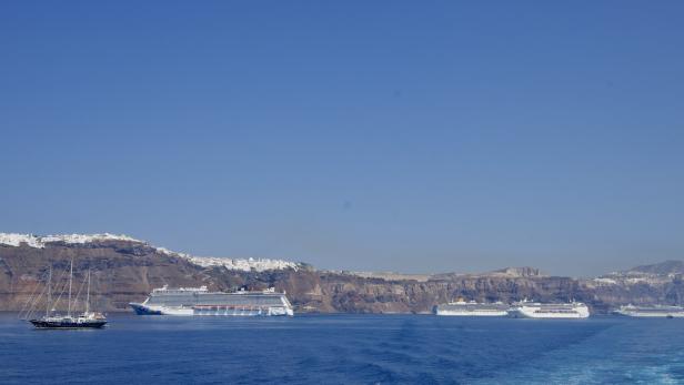 Mehrere Kreuzfahrtschiffe und ein Segelboot liegen vor einer felsigen Küste unter blauem Himmel.