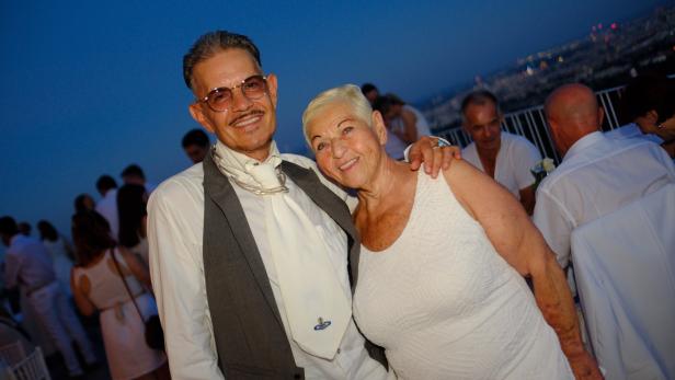 Ein Mann mit Brille und Weste sowie eine Frau in einem weißen Kleid lächeln bei einer Abendveranstaltung in die Kamera.