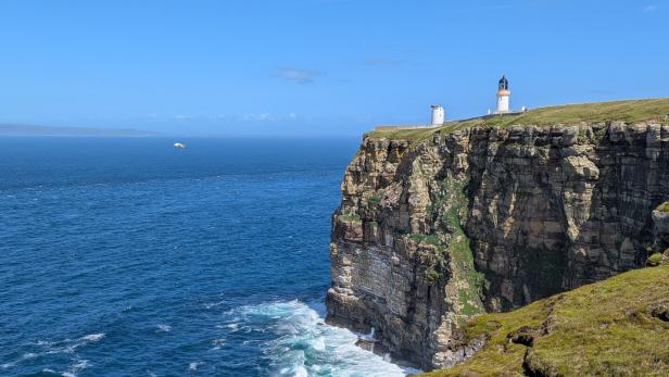 Steilküste am Meer mit Leuchtturm auf der Klippe und Wellenbrandung darunter.