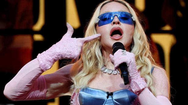 Madonna singt in ein Mikrofon, trägt eine blaue Sonnenbrille, ein blaues Korsett und rosafarbene Spitzenhandschuhe.