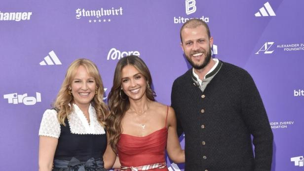 Zwei Frauen und ein Mann posieren vor einer violetten Wand mit Sponsorenlogos, wobei die Frauen in Dirndl und Kleid sowie der Mann in Lederhose auftreten.