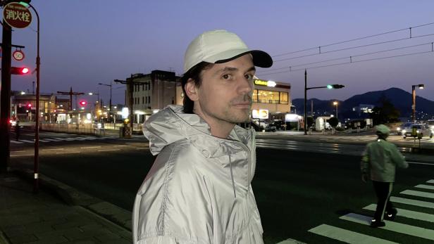 Mann in weißer Kappe und heller Jacke steht an einer nächtlichen Kreuzung mit Ampeln und Bergen im Hintergrund.