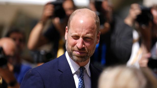 Prinz William trägt einen dunkelblauen Anzug mit Krawatte und blickt vor einer Menschenmenge mit Kameras zur Seite.