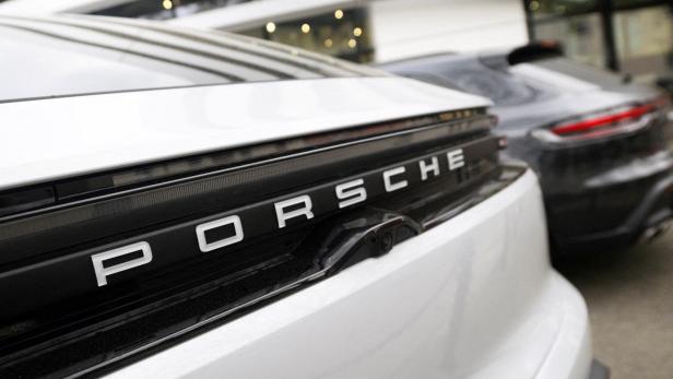Nahaufnahme des markanten Porsche-Schriftzugs am Heck eines weißen Sportwagens vor einem weiteren Fahrzeug.