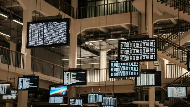 Mehrere Monitore mit Code und Satellitenübersichten hängen in einem großen Innenraum unter einer hohen Decke.