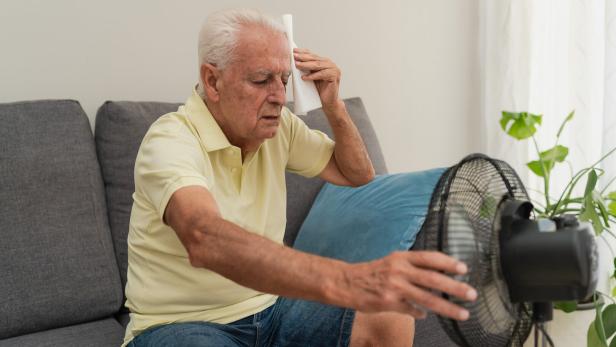 Senior man suffering from summer heat wiping sweat with napkin and holding a electric fan at home