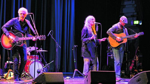 Patti Smith singt auf einer Bühne, begleitet von zwei Gitarristen vor einem blauen Vorhang.