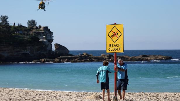 Zwei Rettungsschwimmer stellen am Strand ein Schild mit der Aufschrift „Beach Closed“ auf, während im Hintergrund ein Hubschrauber über dem Meer fliegt.