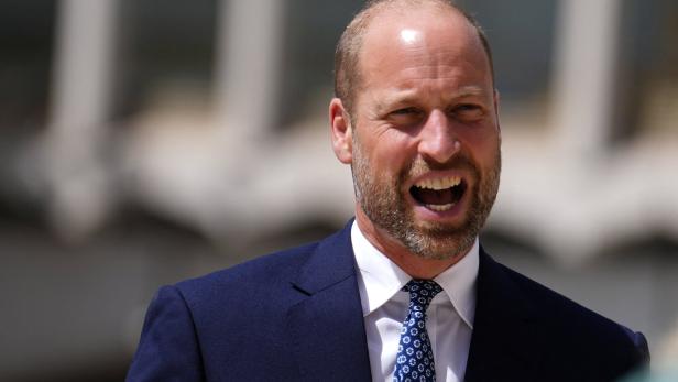 Prinz William trägt einen dunkelblauen Anzug mit weißem Hemd und gemusterter Krawatte und lacht mit offenem Mund.