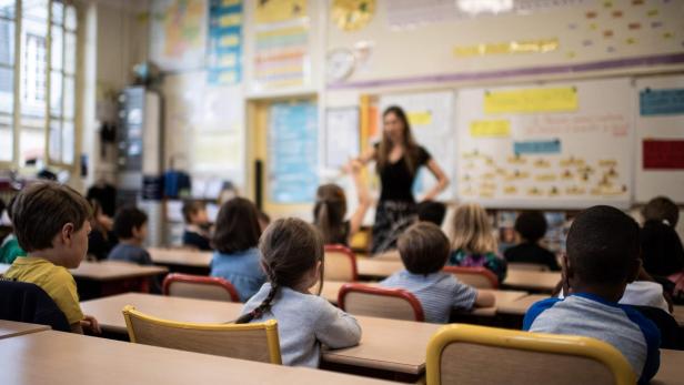 Lehrerin erklärt vor einer Grundschulklasse den Stoff, während die Kinder an Tischen sitzen und zuhören.