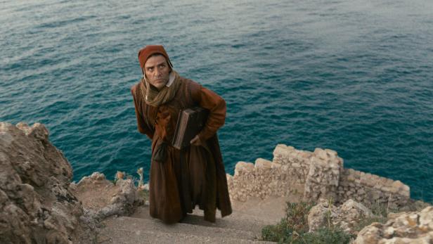 Oscar Isaac in historischer Kleidung trägt ein Buch eine Steintreppe an einer felsigen Küste über dem Meer hinauf.