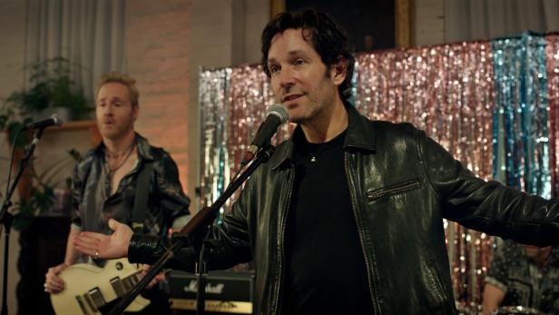 Paul Rudd singt in einer Lederjacke vor einem Mikrofon, während im Hintergrund ein Gitarrist und ein Schlagzeuger auf einer Bühne stehen.