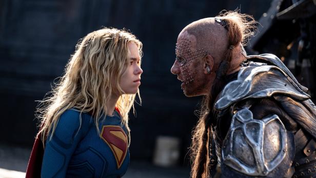 Supergirl steht sich in einem intensiven Blickduell einem kriegerisch gekleideten Gegner mit metallischen Verzierungen im Gesicht gegenüber.