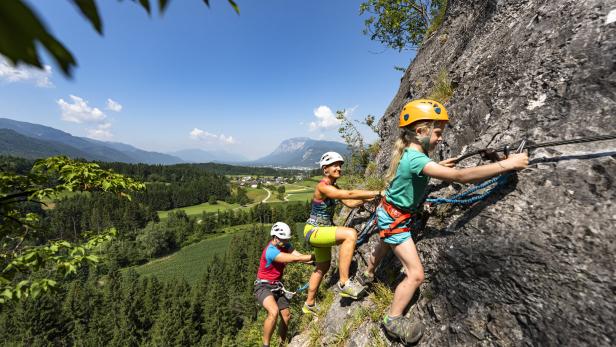 Eine Familie klettert gesichert an einem Felsen mit Blick auf eine grüne Berglandschaft.
