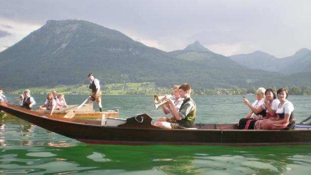 Mehrere Menschen in Tracht fahren und musizieren in Holzbooten auf einem See vor Bergen.