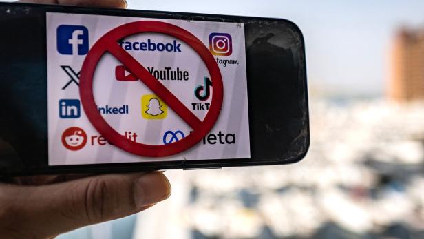 Eine Hand hält ein Smartphone, dessen Display mit einem roten Verbotssymbol über Logos von sozialen Medien wie Facebook, YouTube und TikTok belegt ist.
