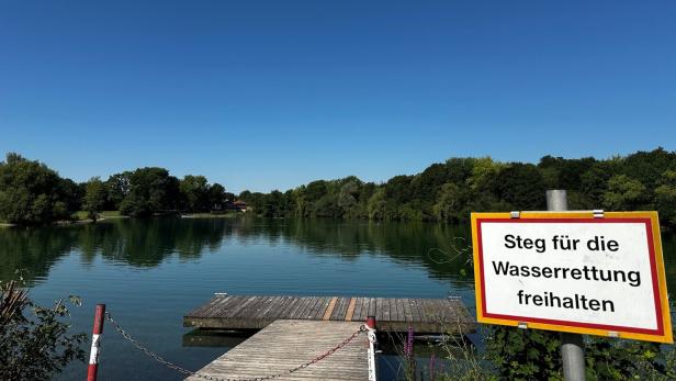 Ruhiger See mit Holzsteg und Schild „Steg für die Wasserrettung freihalten“.