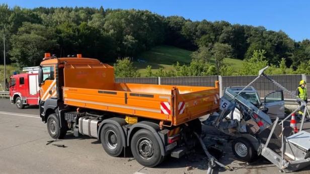Ein orangefarbener Lkw hat einen Kleinwagen mit Anhänger auf der Straße schwer beschädigt, Rettungskräfte stehen daneben.
