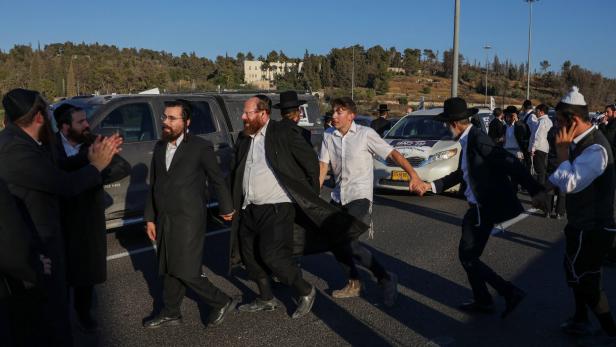 Eine Gruppe orthodoxer jüdischer Männer tanzt Händchen haltend auf einer Straße vor parkenden Autos.