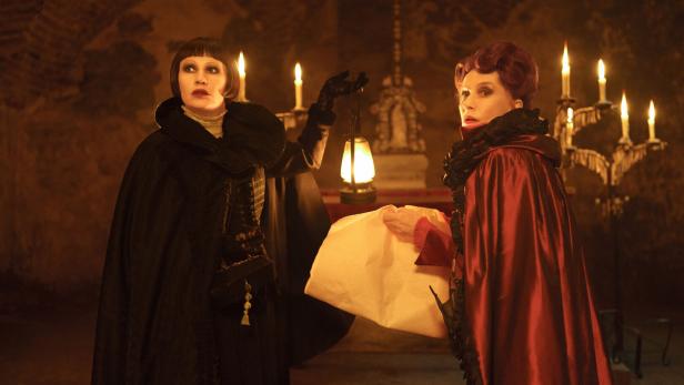 Birgit Minichmayr und Isabelle Huppert in historischen Kostümen und Umhängen stehen in einem dunklen, von Kerzen und einer Laterne beleuchteten Gewölbe.